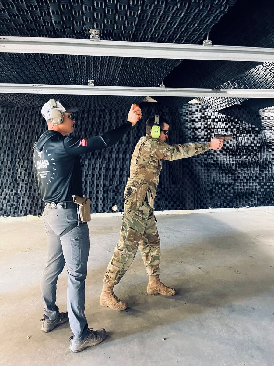 An instructor in a JMAP shirt coaching a service member firing a pistol on an indoor range