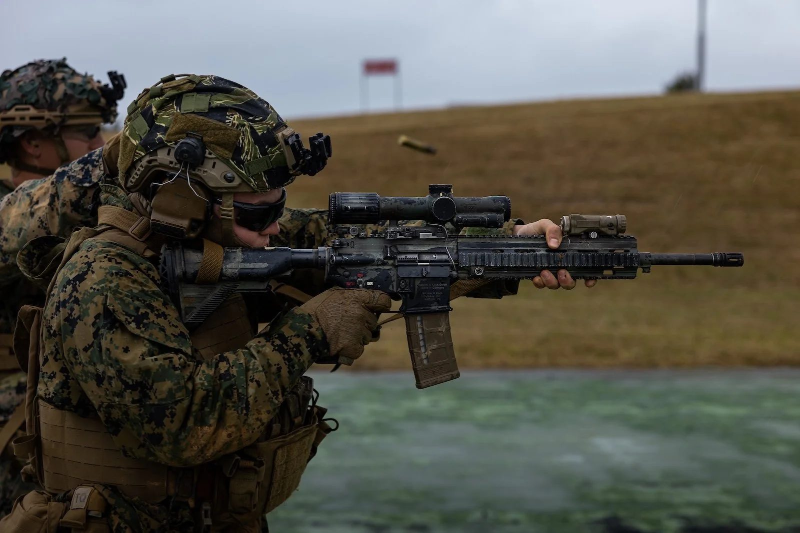 A Marine in a helmet and eye protection aiming a rifle with a magnified optic on a range